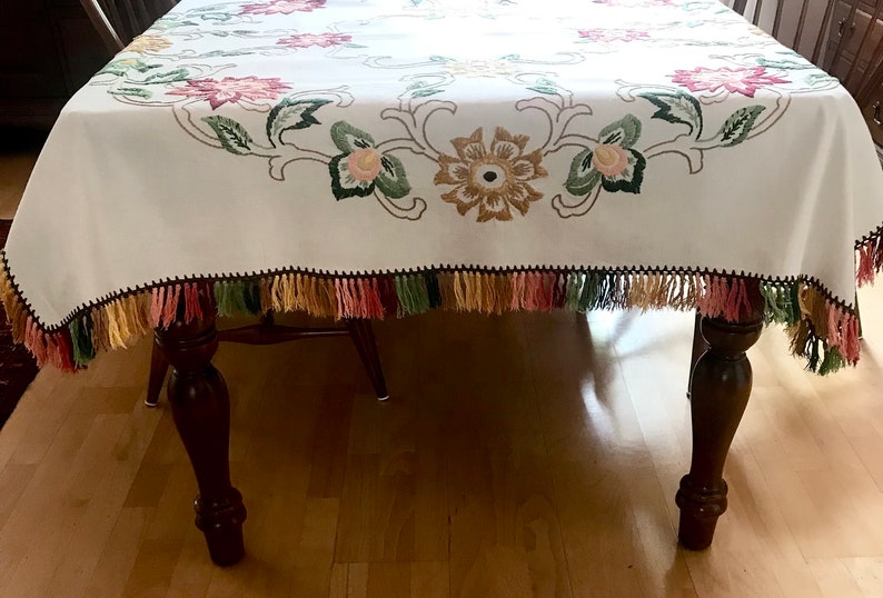May include: White tablecloth with embroidered floral design in pink, green, and gold. The edges are trimmed with a black border and colorful tassels. The tablecloth is draped over a wooden table.