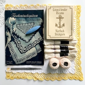 Può includere: Una collezione di forniture vintage per uncinetto e ricamo. Include un libro intitolato "Taschentuchspitzen", una scatola di filo "Coton à broder Ricamo", rocchetti di filo bianco, uncinetti e una bobina blu. Gli articoli sono disposti su una superficie bianca con pizzo giallo e bianco.