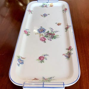 Könnte beinhalten: Ein rechteckiges weißes Serviertablett mit blauem Rand und Blumenmuster. Das Tablett hat an jedem Ende einen Griff und ist mit rosa, blauen und gelben Blumen verziert. Das Tablett ist wahrscheinlich aus Porzellan oder Keramik.