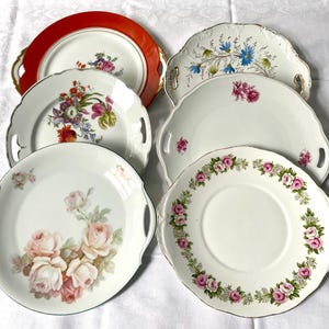 May include: A selection of vintage porcelain serving plates. The plates showcase floral patterns in shades of pink, red, and blue, with gold edging and scalloped rims. Some plates have handles.