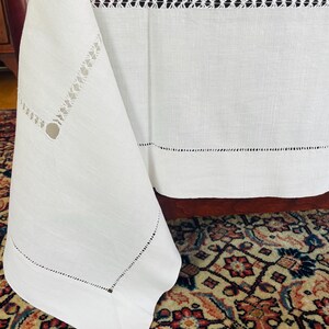 May include: Close-up of a white linen tablecloth with decorative cut-out details. The tablecloth has a textured surface and features a cut-out design along the edges and a horizontal band across the top. The fabric drapes over a wooden surface.