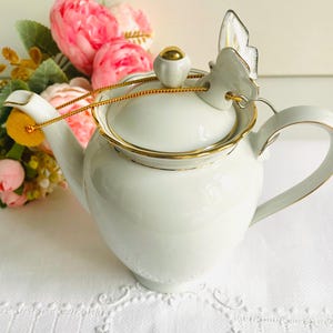 Può includere: Teiera in porcellana bianca con finiture dorate e manico del coperchio a forma di farfalla. La teiera ha un corpo arrotondato e un manico curvo. Una catena dorata è attaccata al coperchio. Fiori rosa e gialli sullo sfondo.