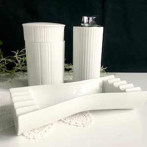 Pode incluir: Cinzeiro, isqueiro e pote de cerâmica branca com design canelado. O cinzeiro é curvo e tem uma forma longa e estreita.