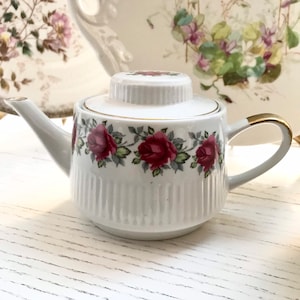 Può includere: Teiera bianca con bordo dorato e un motivo di rose rosse. La teiera ha un design a coste e un coperchio.