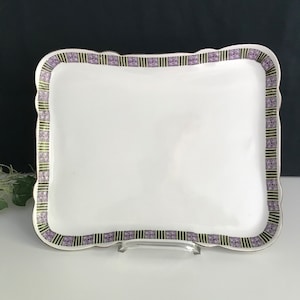 Può includere: Un vassoio da portata quadrato bianco con un bordo smerlato. Il vassoio ha un bordo decorativo di quadrati viola e verdi.