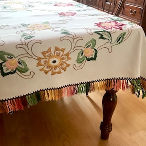 May include: Cream-colored tablecloth with embroidered floral design in shades of yellow, pink, and green. The edges are trimmed with a fringe in matching colors. The tablecloth is draped over a wooden table.