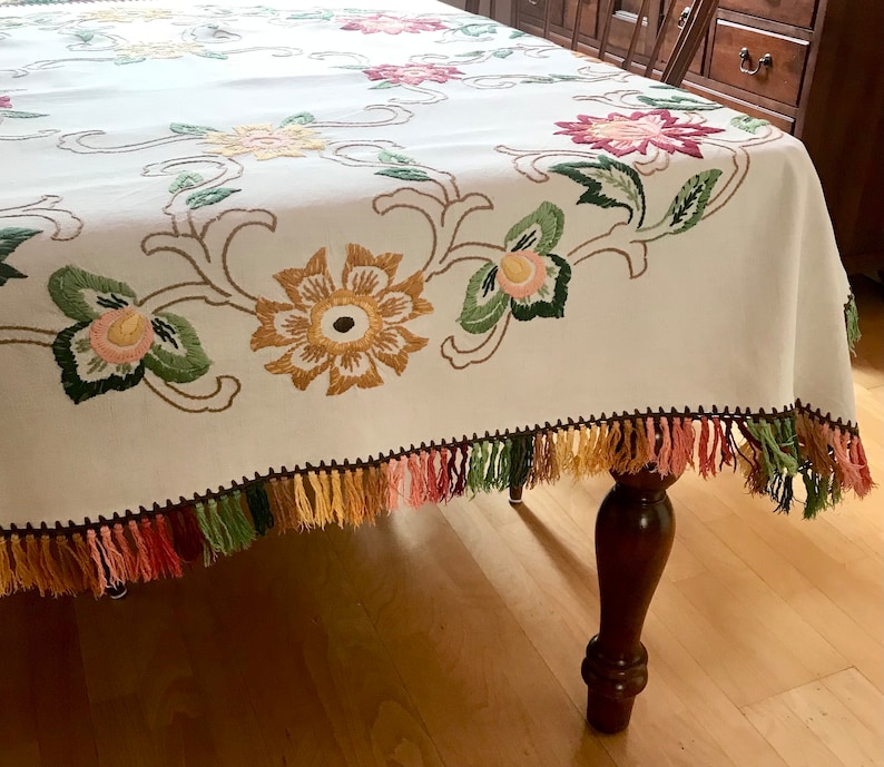 May include: Cream-colored tablecloth with embroidered floral design in shades of yellow, pink, and green. The edges are trimmed with a fringe in matching colors. The tablecloth is draped over a wooden table.