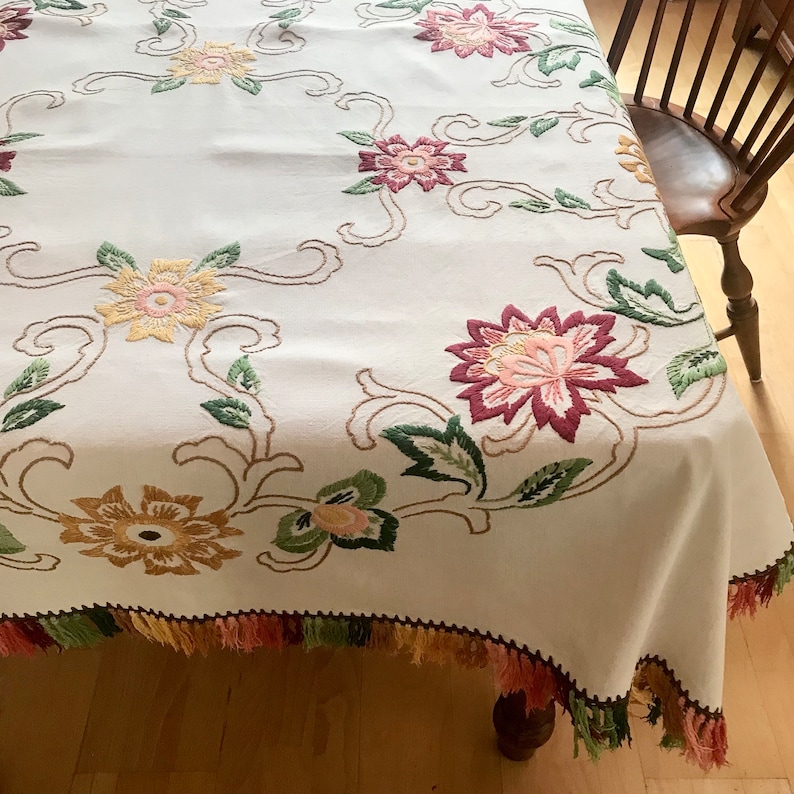 May include: Cream-colored tablecloth with embroidered floral design in shades of pink, yellow, and green. The edges are trimmed with a colorful fringe. The tablecloth is draped over a wooden table, showcasing the intricate details of the embroidery.