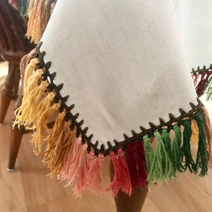 May include: A white linen tablecloth with a dark brown crocheted edge and colorful fringe. The fringe includes shades of yellow, orange, pink, red, green, and brown. The tablecloth is draped over a wooden table.