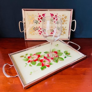 Könnte beinhalten: Zwei rechteckige Serviertabletts mit Metallrahmen und Griffen, die cremefarbene Oberflächen mit Blumenmustern in Rot, Grün und Gold aufweisen. Ein klares Weinglas aus Glas steht auf dem vorderen Tablett, das ein großes rotes Blumenmuster zeigt.