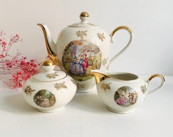 May include: A vintage porcelain tea set with gold accents. The teapot, sugar bowl, and creamer feature a delicate floral design and a romantic scene with figures in a garden.