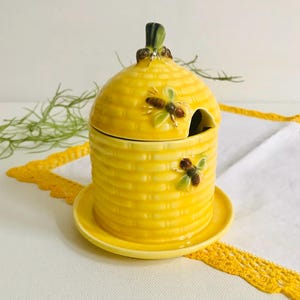 Può includere: Un vasetto di miele in ceramica gialla a forma di alveare, con coperchio e un cucchiaino. Il vasetto ha una superficie strutturata ed è decorato con due figurine di api. Poggia su un piattino abbinato.