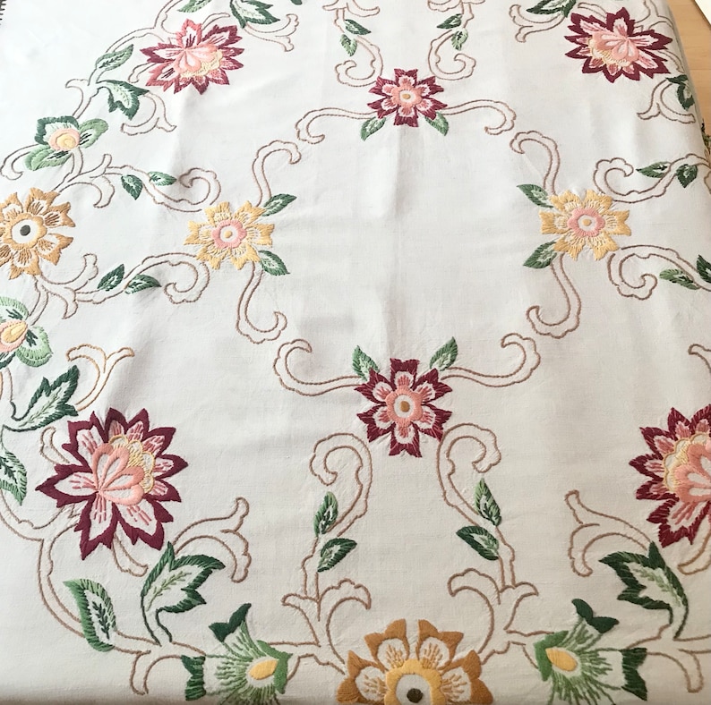 May include: Cream-colored embroidered tablecloth featuring floral patterns in shades of pink, yellow, and burgundy. The design includes detailed flower and leaf motifs with a delicate, flowing aesthetic. The embroidery is done with a variety of stitches.