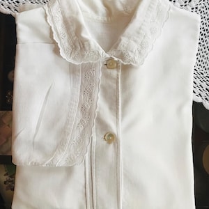 May include: White cotton shirt with a lace collar and trim. The shirt has a button-up front and long sleeves.