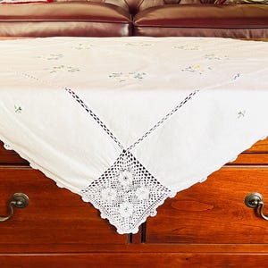 Könnte beinhalten: Eine quadratische weiße Tischdecke mit zarter Blumenstickerei in sanften Pastelltönen. Die Kanten sind mit aufwendigen weißen Spitzendetails und einem gewellten Rand versehen, was ein dekoratives Textilstück für eine Tischoberfläche ergibt.