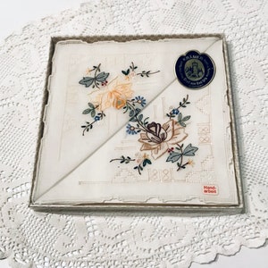 Könnte beinhalten: Zwei weiße Taschentücher mit zarter Blumenstickerei in Rosa-, Gelb- und Grüntönen. Die Taschentücher befinden sich in einer Schachtel mit dem Text "Hand-arbeit" und einem Etikett mit dem Text "KRÄMER" und einer Schlossgrafik.