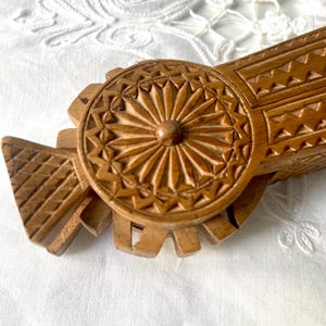 Op de afbeelding: Een close-up van een vintage houten notenkraker. De notenkraker is gesneden uit lichtbruin hout en heeft een cirkelvormig, stervormig patroon. Het ontwerp is omgeven door geometrische vormen en lijnen.