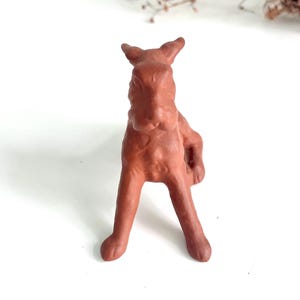Könnte beinhalten: Eine terrakottafarbene Keramik-Hasenfigur. Der Hase ist in sitzender Position mit ausgestreckten Vorderpfoten dargestellt. Die Ohren sind aufrecht, und das Gesamtdesign ist schlicht und rustikal. Die Figur steht auf einer weißen Oberfläche.