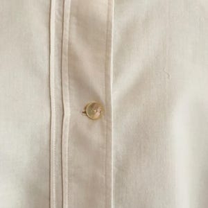 May include: A close-up of a white button-down shirt with a single button fastened at the centre. The shirt has a crisp, clean look and is made of a smooth, lightweight fabric.