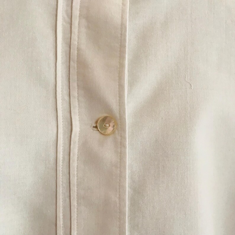 May include: A close-up of a white button-down shirt with a single button fastened at the centre. The shirt has a crisp, clean look and is made of a smooth, lightweight fabric.