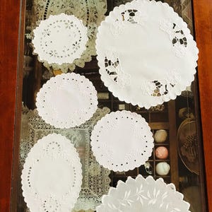 Può includere: Una collezione di sette tovagliette all'uncinetto bianche con disegni intricati, esposte su una superficie di vetro con una cornice in legno.