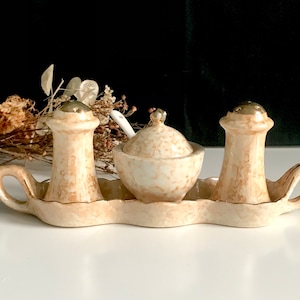 Könnte beinhalten: Ein Set aus drei Keramik-Salz- und Pfefferstreuern und einer Zuckerdose mit passendem Tablett. Die Streuer und die Dose haben eine gesprenkelte beige und weiße Glasur mit goldenen Akzenten.