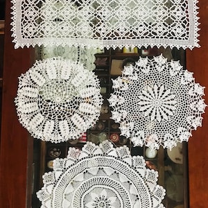May include: Four white crocheted doilies, two rectangular and two round, displayed on a wooden surface.