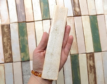 May include: A single white ceramic tile with a rough texture. The tile is approximately 6 inches long and 1.5 inches wide. The tile is held in a hand against a background of similar tiles in various colors and patterns.
