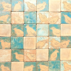 Könnte beinhalten: Eine Sammlung von 25 Keramikfliesen mit einer blauen und beigefarbenen Glasur. Jede Fliese zeigt ein handgemaltes Bild eines Vogels in einem einfachen, stilisierten Design.