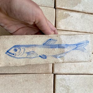 Könnte beinhalten: Eine rechteckige, cremefarbene Fliese mit einem handgezeichneten blauen Fischdesign. Der Fisch ist in einem einfachen Stil dargestellt, mit sichtbaren Details wie Flossen und Schuppen. Die Fliese wird von einer Hand gehalten, im Hintergrund sind weitere ähnliche Fliesen zu sehen.