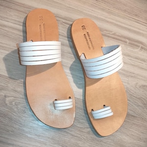May include: A pair of white leather sandals with a toe thong and multiple straps. The sandals are size 38 and have the text "HAND FINISHED -  MADE IN GREECE - GENUINE LEATHER" on the straps.
