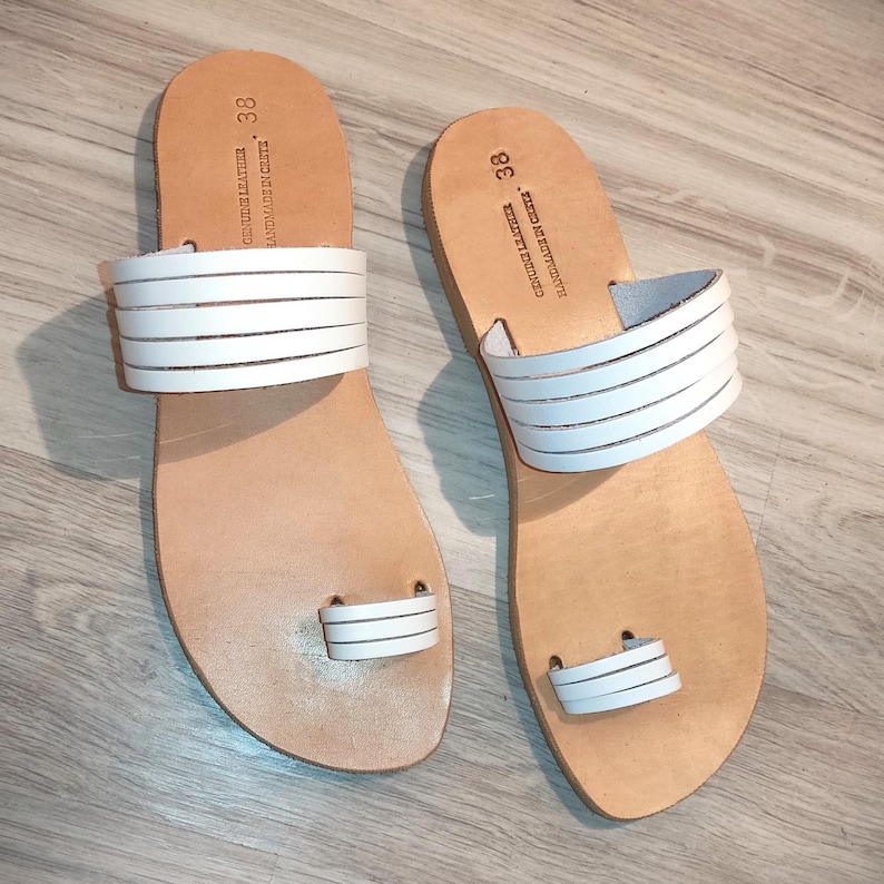 May include: A pair of white leather sandals with a toe thong and multiple straps. The sandals are size 38 and have the text "HAND FINISHED -  MADE IN GREECE - GENUINE LEATHER" on the straps.