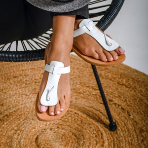May include: White leather sandals with a buckle strap and a brown sole. The sandals are worn by a person sitting on a black chair with a woven straw rug underneath.