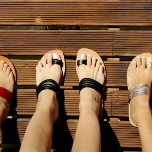 May include: Five pairs of leather sandals with different colored straps. The sandals are all brown leather with a toe loop and a strap across the foot. The straps are black, red, silver, gold, and white.