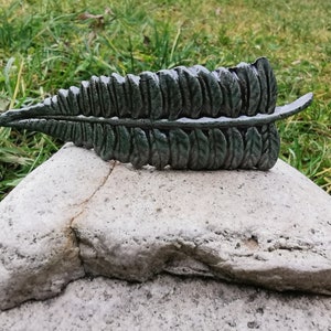 Könnte beinhalten: Eine Keramik-Skulptur in Form eines Farnblattes in dunkelgrüner Farbe. Das Blatt ist mit einzelnen Blättchen detailliert und hat eine strukturierte Oberfläche.