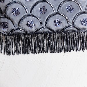 Puede incluir: Un bolso de mano gris con cuentas y un borde de flecos. El bolso est&aacute; decorado con un patr&oacute;n de lentejuelas y cuentas en forma de escamas.