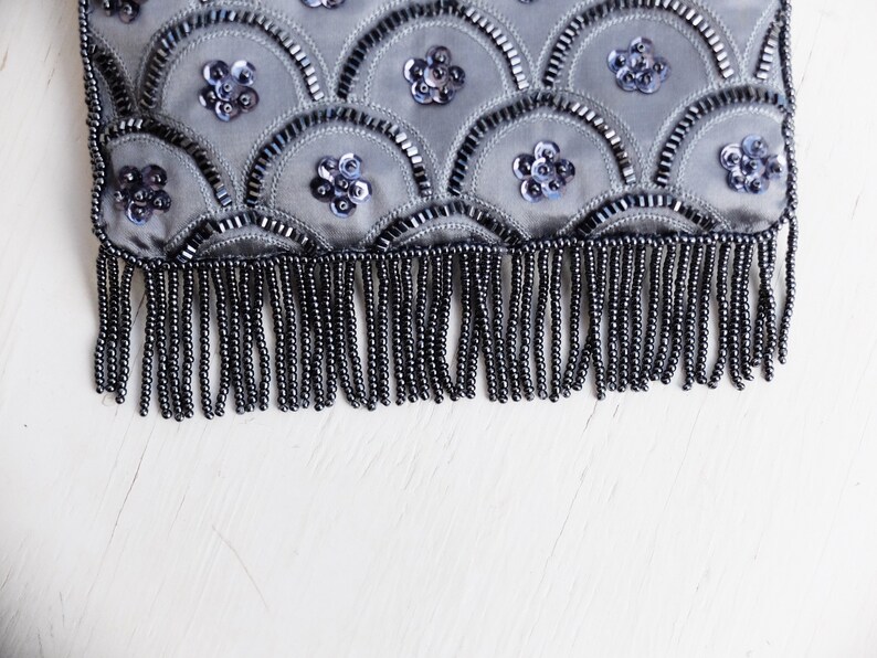 Puede incluir: Un bolso de mano gris con cuentas y un borde de flecos. El bolso est&aacute; decorado con un patr&oacute;n de lentejuelas y cuentas en forma de escamas.