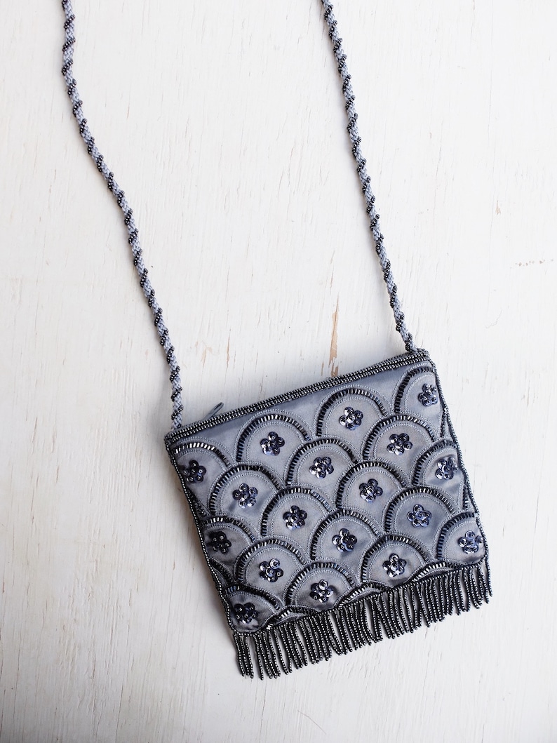 Puede incluir: Un bolso de noche gris con cuentas, con una correa de cadena plateada y un dise&ntilde;o de concha. El bolso tiene un ribete de flecos.
