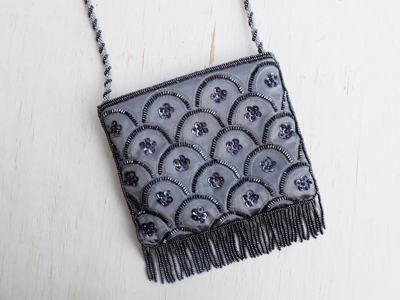 Puede incluir: Un bolso de noche gris con cuentas y un detalle de flecos. El bolso est&aacute; decorado con un patr&oacute;n de lentejuelas y cuentas.