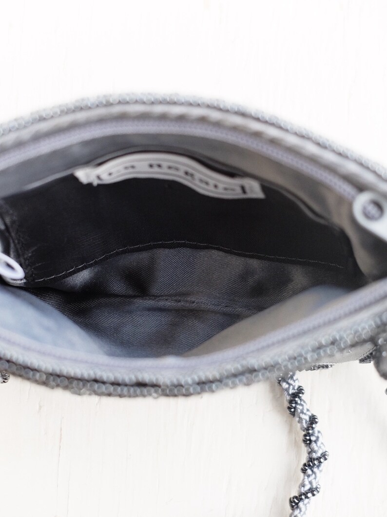 Puede incluir: Un bolso de noche gris con cuentas, con cierre de cremallera y una correa de cuentas negras y grises. El bolso tiene un forro de sat&eacute;n negro y una peque&ntilde;a etiqueta blanca en el interior que dice "La Regale".