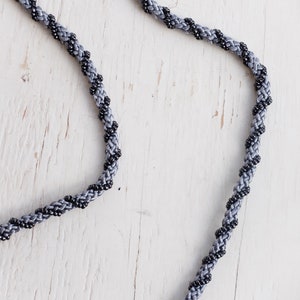 Puede incluir: Un collar de cord&oacute;n trenzado gris y negro con un dise&ntilde;o trenzado.