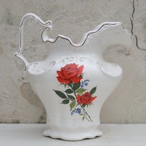 May include: White ceramic pitcher with a decorative handle and floral design. The pitcher features red roses and blue flowers.