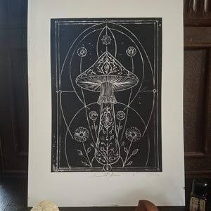 Könnte beinhalten: Ein schwarz-weißer Linoldruck auf Papier, der einen zentralen Pilz mit komplizierten floralen und geometrischen Mustern zeigt. Das Kunstwerk ist von einem bogenförmigen Rand umgeben und enthält den handschriftlichen Text Amanita Arcana unten.