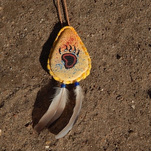 Puede incluir: Un colgante de cuero amarillo con una huella de pata de oso pintada y detalles en azul y rojo. El colgante está unido a un cordón de cuero marrón y tiene dos plumas blancas colgando.