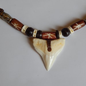 Op de afbeelding: Een bruine en witte kralenketting met een witte haaientand hanger. De kralen zijn gemaakt van hout en hebben een stervormig ontwerp.