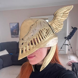 K&ouml;nnte beinhalten: Ein goldfarbener Helm mit filigranen Details und einem Fl&uuml;geldesign. Der Helm ist aus leichtem Material gefertigt und hat einen schlanken, modernen Look.