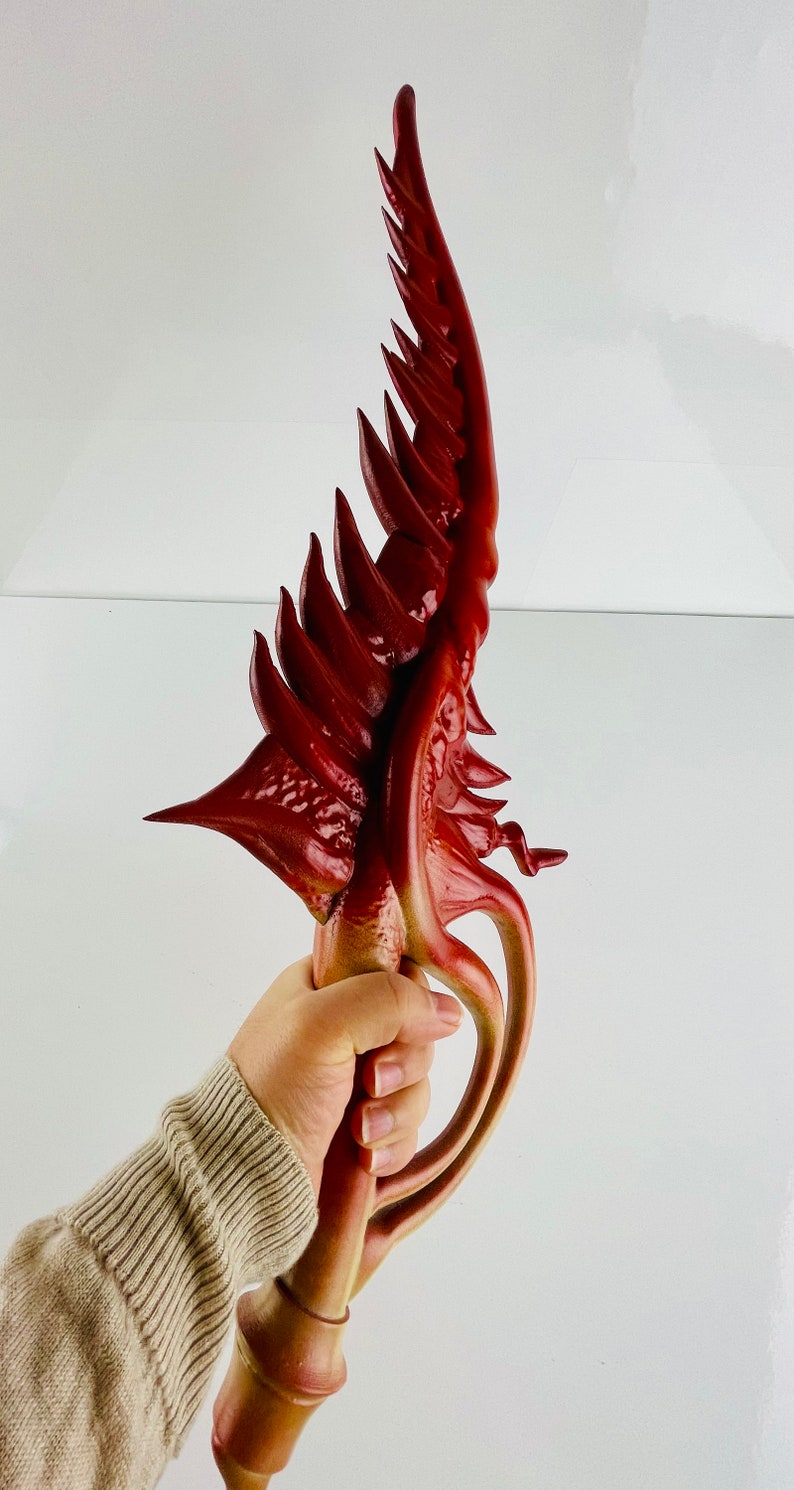 May include: A red and gold decorative sword with a wing-like design. The sword is held in a hand.