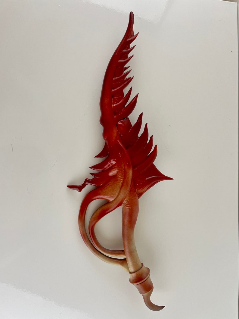 May include: A red and orange decorative sword with a curved blade and a pointed tip. The blade is decorated with intricate details and has a textured surface.