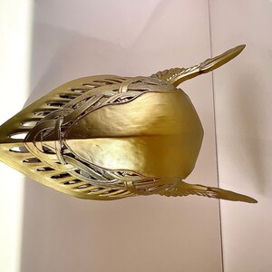 K&ouml;nnte beinhalten: Ein goldfarbener Helm mit filigranen Details und einer spitzen Oberseite. Der Helm hat ein fl&uuml;gelartiges Design an den Seiten.