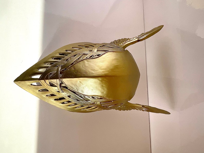 K&ouml;nnte beinhalten: Ein goldfarbener Helm mit filigranen Details und einer spitzen Oberseite. Der Helm hat ein fl&uuml;gelartiges Design an den Seiten.